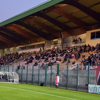Spectateurs assis dans la tribune couverte du stade Pierre-Brisson de Beauvais, un soir de match de football sous les projecteurs, avec un panneau publicitaire le long de la main courante