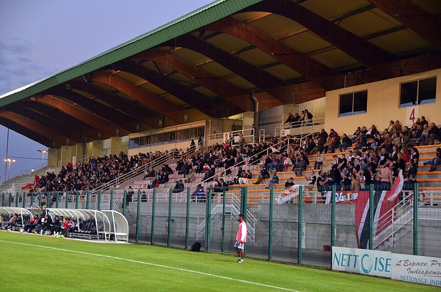 Spectateurs assis dans la tribune couverte du stade Pierre-Brisson de Beauvais, un soir de match de football sous les projecteurs, avec un panneau publicitaire le long de la main courante