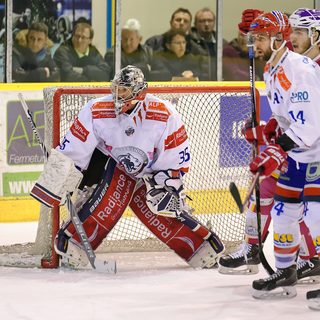 Un gardien de hockey sur glace posté devant sa cage pendant un match de Ligue Magnus, deux joueurs adverses devant lui et le public installé derrière les balustrades