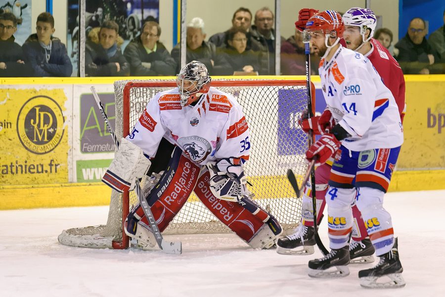 Un gardien de hockey sur glace posté devant sa cage pendant un match de Ligue Magnus, deux joueurs adverses devant lui et le public installé derrière les balustrades