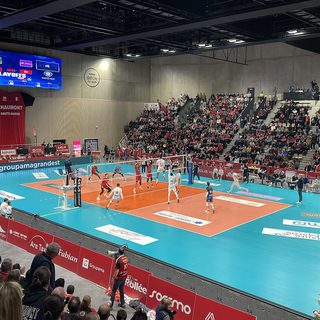Vue d'ensemble d'une salle de volley-ball pendant un match de playoffs du championnat de France, le terrain bleu et orange au centre et les tribunes garnies de supporters