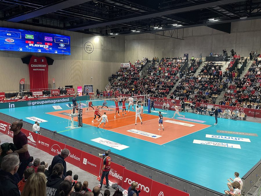 Vue d'ensemble d'une salle de volley-ball pendant un match de playoffs du championnat de France, le terrain bleu et orange au centre et les tribunes garnies de supporters