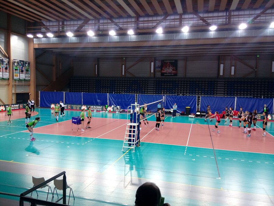 L'intérieur de la salle Maréchal de Harnes avant un match de volley, les deux équipes à l'échauffement sur le terrain devant les tribunes
